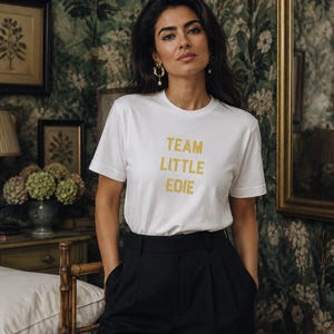 South Asian woman modelling a white Team Little Edie graphic T-shirt with black tailored trousers against dark botanical wallpaper in an editorial interior.