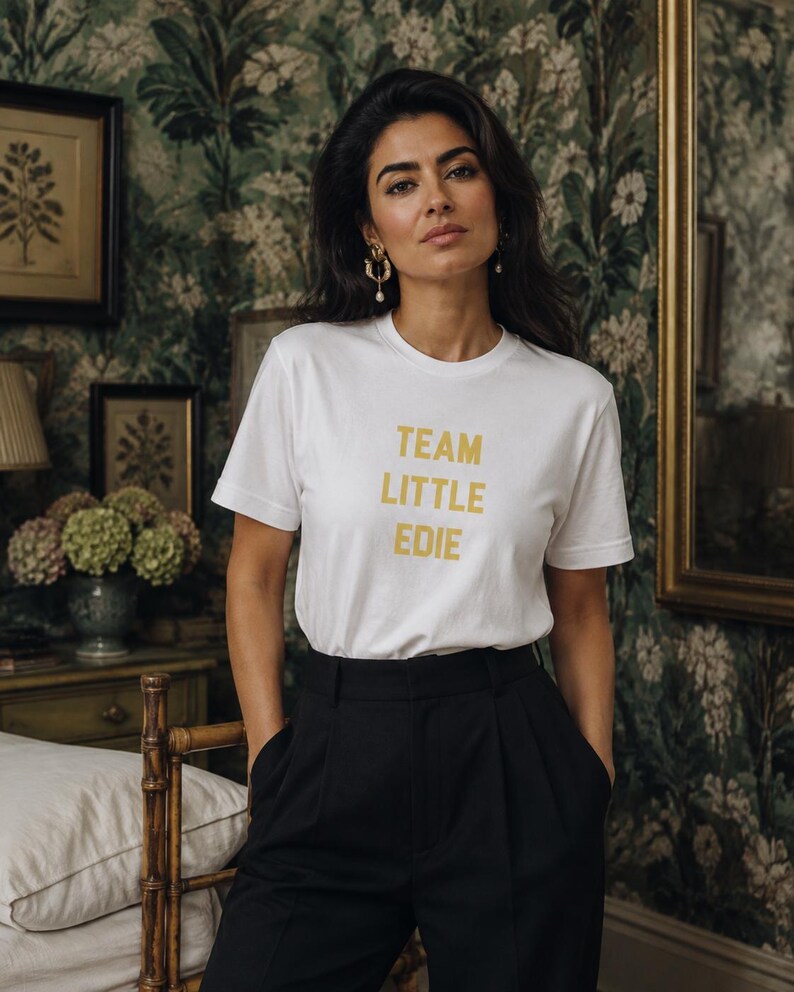 South Asian woman modelling a white Team Little Edie graphic T-shirt with black tailored trousers against dark botanical wallpaper in an editorial interior.