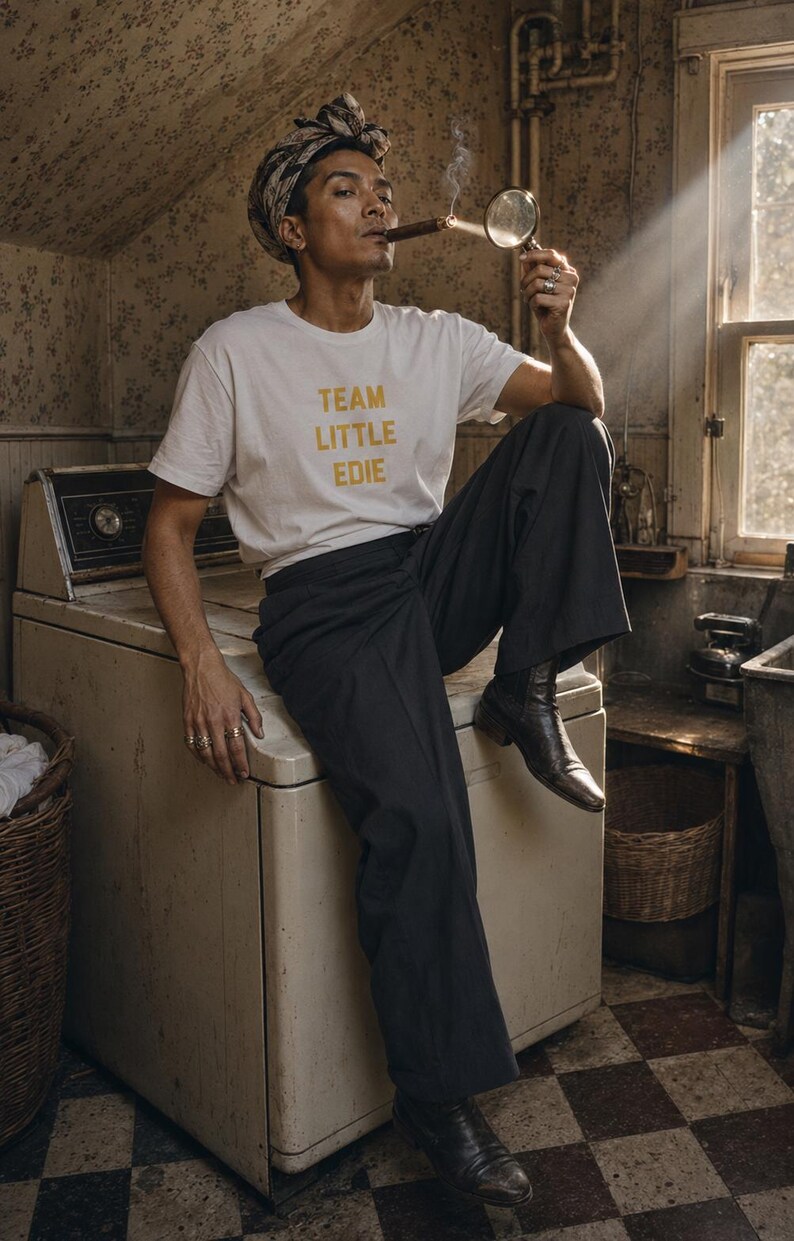 Non-binary adult wearing a white Team Little Edie T-shirt and patterned headscarf, seated on a vintage washing machine while lighting a cigar with sunlight and a magnifying glass.