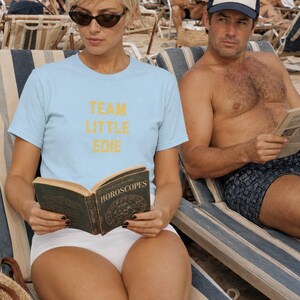 Blonde woman wearing a pale blue Team Little Edie T-shirt and reading a horoscope book on a crowded beach beside a man wearing a Libra Man trucker hat.