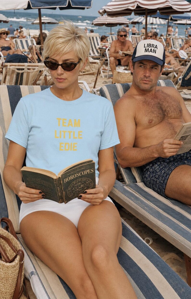 Blonde woman wearing a pale blue Team Little Edie T-shirt and reading a horoscope book on a crowded beach beside a man wearing a Libra Man trucker hat.
