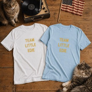 White and pale blue Team Little Edie graphic T-shirts arranged on aged oak floorboards with two long-haired cats, a vintage record player and a small handheld American flag.