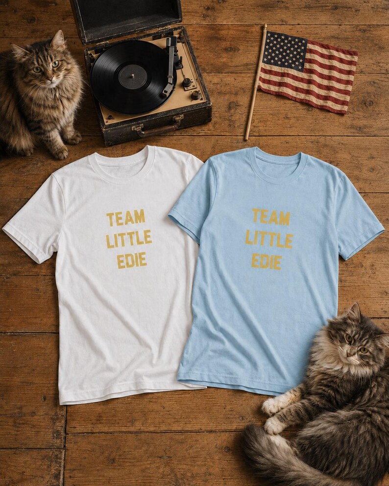 White and pale blue Team Little Edie graphic T-shirts arranged on aged oak floorboards with two long-haired cats, a vintage record player and a small handheld American flag.