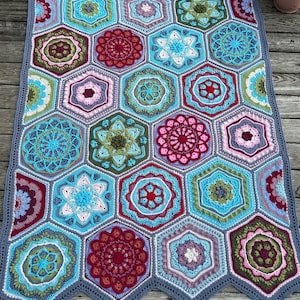 Puede incluir: Una manta de ganchillo con un patrón hexagonal en tonos de azul, verde, rosa y rojo. La manta está compuesta por hexágonos individuales, cada uno con un diseño diferente. La manta está bordeada con un ribete de ganchillo gris.