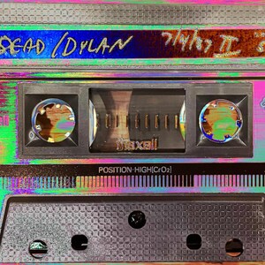 May include: A vintage cassette tape labeled "DEAD /DYLAN" with the text "기어" and "S74" on the label. The tape is a Maxell Epitaxial XLII 90, made in Japan.
