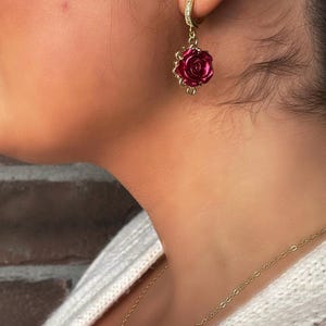 May include: Gold hoop earrings with a dangling red rose charm. The rose is made of a shiny, textured material.