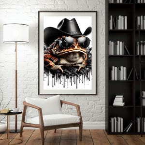 May include: A framed art print featuring a close-up of a toad wearing a black cowboy hat and sunglasses. The toad has brown and orange skin. Black paint drips down from the bottom of the image.