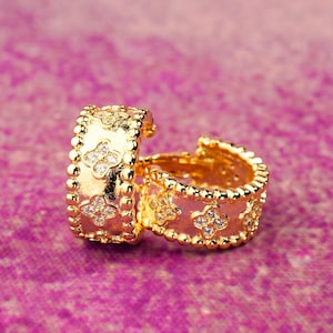 Könnte beinhalten: Ein Paar goldfarbene Creolen mit floralem Design und kleinen, klaren Edelsteinen. Die Ohrringe haben einen strukturierten, perlenbesetzten Rand und sind vor einem rosa Hintergrund platziert.