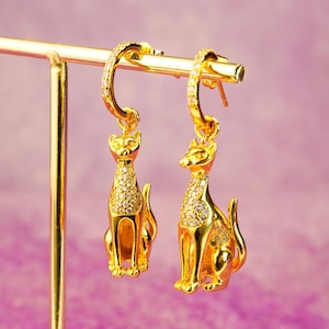 Op de afbeelding: Een paar gouden creolen met hangende kattenbedels. De katten zijn versierd met glinsterende, heldere strasssteentjes.