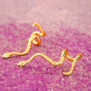 Könnte beinhalten: Ein Paar goldene Schlangenohrringe mit klaren Kristall-Akzenten. Die Ohrringe sind so konzipiert, dass sie sich um das Ohrläppchen wickeln.