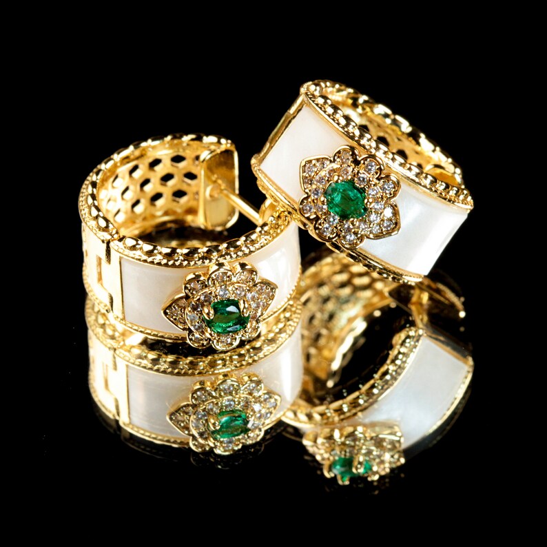 May include: A pair of gold hoop earrings with white enamel and green gemstones. The earrings are set with small diamonds and have a floral design.