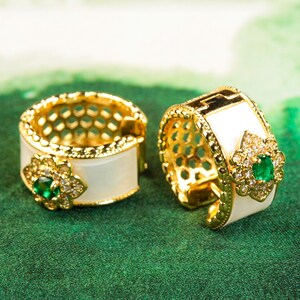 May include: A pair of gold hoop earrings with white enamel and green gemstone accents. The earrings feature a delicate floral design.