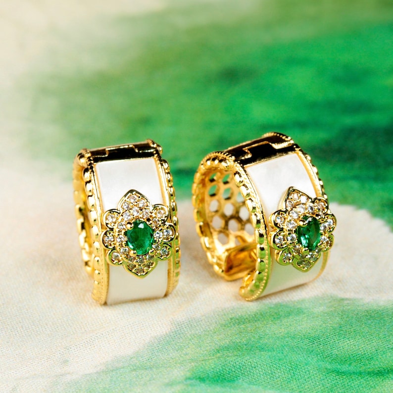 May include: A pair of gold hoop earrings with white enamel and a green gemstone surrounded by clear rhinestones.