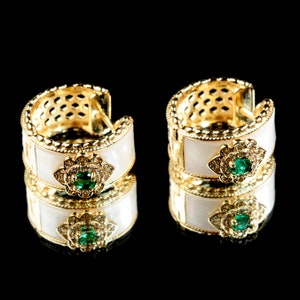 May include: A pair of gold hoop earrings with white enamel and green gemstones. The earrings are reflected in a black surface.
