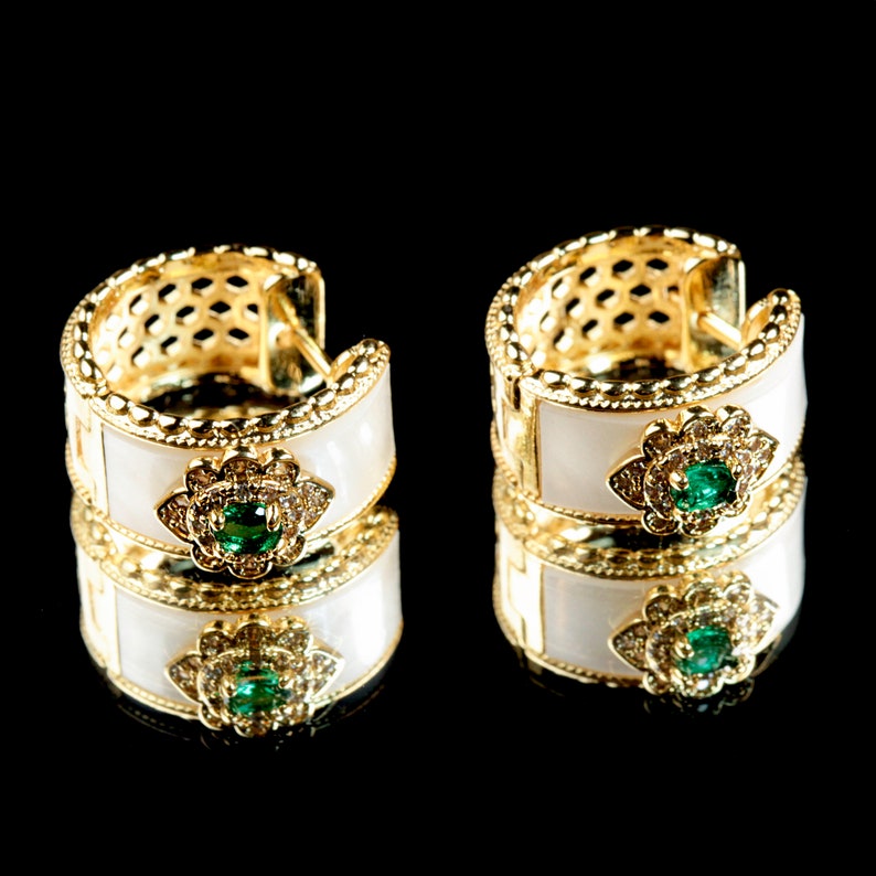 May include: A pair of gold hoop earrings with white enamel and green gemstones. The earrings are reflected in a black surface.