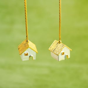 May include: Two miniature silver houses with gold roofs hang from gold chains against a green background.