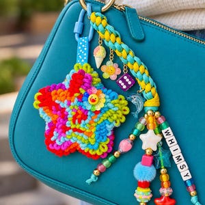 May include: A multi-coloured crochet star keyring attached to a teal handbag. The accessory features a braided teal and yellow cord, dangling charms including a miniature ice cream cone, a flower, a purple die, a dolphin, and beaded strands with the word WHIMSY.