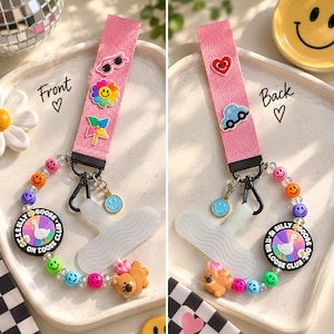 May include: A pink fabric wristlet strap featuring colorful charms including sunglasses, a flower, and a pinwheel. Attached is a beaded loop with a round charm reading Silly Goose On Loose Club, a white wavy clip, and a brown bear charm.
