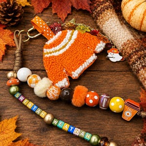 Könnte beinhalten: Ein Perlen-Schlüsselanhänger mit einem orangefarbenen Pullover-Anhänger in Punch-Needle-Technik, einem Geist und einem Kaffeebecher mit der Aufschrift FALL FUN. Die Perlen sind orange, weiß, grün und Buchstabenwürfel, die SWEATAH WEATHAH bilden.