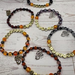 May include: Five beaded bracelets featuring round beads in various colours including black, orange, red, and purple. Each bracelet includes gold-toned letter beads spelling out RIDER, STORM, BASGIATH, ANDARNA, VIOLENCE, and FLAME, along with metal dragon charms.
