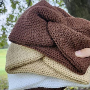 May include: A stack of three knitted headbands in shades of brown, tan, and white. The headbands are made of a textured knit fabric and have a twisted knot design. The headbands are soft and cozy, perfect for keeping ears warm.