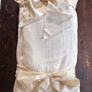 May include: Cream-colored, textured fabric item with a grid pattern, tied with silk bows. The bows are a lighter shade of cream and are tied around the center and top of the item. The item appears to be a blanket or wrap.