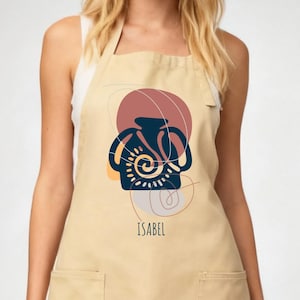 May include: Beige apron with two front pockets and a neck strap. The apron features a navy blue and yellow abstract design of a vase with a spiral pattern, set against a maroon circle and the name "ISABEL" printed below.