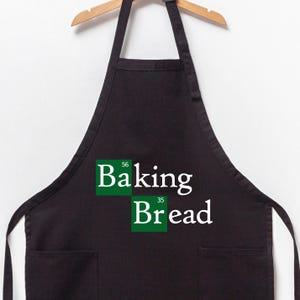 May include: Black apron with a white pocket and a green and white graphic that says "Baking Bread" with the chemical element symbols for baking soda and flour.