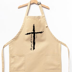 May include: Beige apron with two front pockets and a wooden hanger. A large, black, brushstroke-style cross is centered on the apron. The text "Use your gift to serve" is written vertically on the left side of the cross, with "1 Peter 4:10" on the right.
