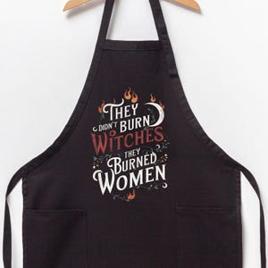 May include: Black apron with two front pockets and a wooden hanger. The apron features the text "They didn't burn witches, they burned women" in white and red lettering, with decorative floral and flame accents.