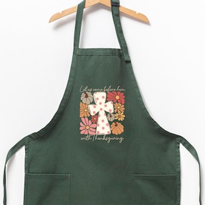 May include: A dark green apron with two front pockets, hanging on a wooden hanger. The apron features a floral design with a cross and the text "Let us come before him with Thanksgiving."
