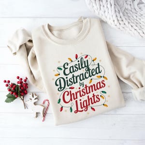 May include: A beige sweatshirt with the text "Easily Distracted by Christmas Lights" in green and red, decorated with string lights. A sprig of red berries, a wooden reindeer, and a candy cane are on the left.