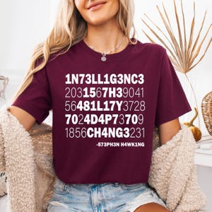 May include: A maroon t-shirt with white text that reads "1N73LL1G3NC3 2031567H39041 56481L17Y3728 7024D4P73709 1856CH4NG3231 -573PH3N H4WK1NG"