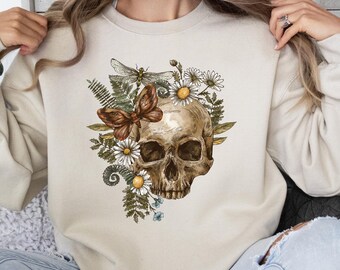 May include: Beige sweatshirt featuring a detailed illustration of a human skull adorned with daisies, ferns, a dragonfly, and a butterfly. The design has a vintage, botanical aesthetic, with a focus on natural elements and a touch of gothic style.