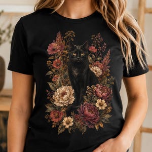 May include: A black short-sleeved t-shirt featuring a graphic print of a black cat sitting amidst a cluster of colourful flowers in shades of red, pink, and cream. The cat has yellow eyes and is positioned centrally on the front of the shirt.
