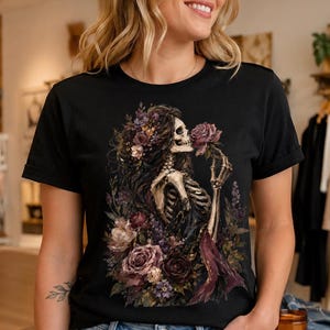 May include: A black t-shirt featuring a graphic print of a skeleton with long dark hair, surrounded by a cluster of deep purple, pink, and dark red roses and foliage. The skeleton is holding a single rose near its face in a detailed illustration.