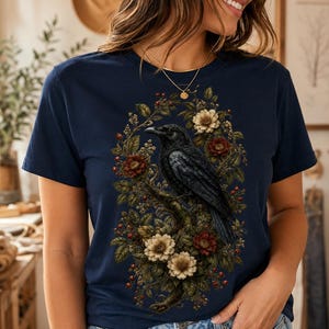 May include: A navy blue short-sleeve t-shirt featuring a detailed graphic print of a black raven perched on a branch, surrounded by a lush arrangement of dark red and cream-colored flowers, green leaves, and small red berries in an oval shape.