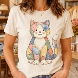 May include: A cream-colored short-sleeve t-shirt featuring a large graphic of a sitting cat made of various quilted fabric patterns. The cat design includes patches of blue gingham, pink, yellow, and floral prints with visible stitched outlines.