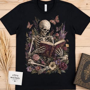 Skeleton reading book surrounded by flowers and butterflies on black t-shirt.