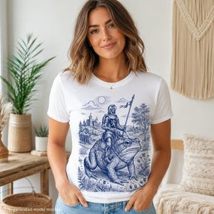May include: A white short-sleeve t-shirt featuring a blue ink-style graphic print of a knight in full armor riding a large toad. The illustration includes a castle in the background, a flag held by the knight, trees, and various plants.
