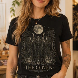 May include: A black short-sleeve t-shirt featuring a graphic print of three cloaked figures standing beneath a full moon, surrounded by trees and crystals, with the text THE COVEN printed in a serif font at the bottom of the design.