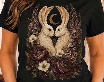 May include: A black t-shirt featuring a graphic print of two cream-colored rabbits facing each other, surrounded by dark purple and white flowers, green foliage, and a golden crescent moon positioned between the rabbits' ears.