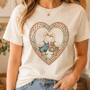 以下が含まれることがあります： クリーム色の半袖Tシャツで、中央にハート型のフレームに入った2羽のアヒルのグラフィックが描かれています。アヒルは服を着ており、ハートの縁はカラフルで複雑なキルト風の花柄で装飾されています。