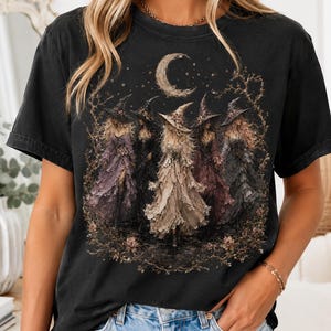 May include: A black short-sleeve t-shirt featuring a graphic print of five figures wearing pointed hats and textured cloaks standing under a crescent moon and stars. The design includes muted tones of beige, brown, and purple within the illustration.