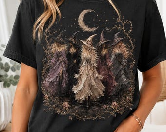 May include: A black short-sleeve t-shirt featuring a graphic print of five figures wearing pointed hats and textured cloaks standing under a crescent moon and stars. The design includes muted tones of beige, brown, and purple within the illustration.