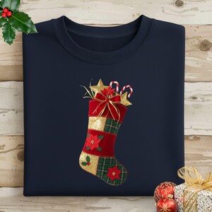 Puede incluir: Sudadera azul marino con un diseño de calcetín navideño. El calcetín es rojo, dorado y verde, con una estrella, bastones de caramelo y un lazo rojo. Las flores de Pascua y el acebo adornan el calcetín.