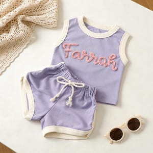 May include: A lavender tank top and matching shorts set with cream-colored trim. The tank top features the name "Farrah" in pink embroidered letters. The shorts have a drawstring waist and cream-colored trim. Sunglasses are also in the image.