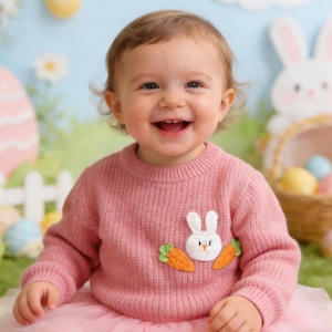 May include: A smiling child in a pink sweater featuring a white bunny and carrot design, paired with a pink tutu. The background includes Easter decorations, such as a basket with eggs and a large Easter egg.