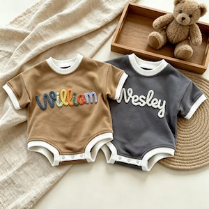 May include: Two baby bodysuits. One is brown with the name "William" embroidered in colorful thread. The other is gray with the name "Wesley" embroidered in white. Both have white trim around the neck, sleeves, and legs.