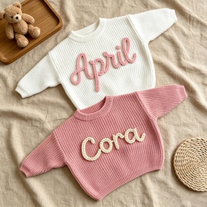 May include: Two long-sleeved ribbed knit sweaters. The top sweater is white with the name April embroidered in pink yarn. The bottom sweater is dusty pink with the name Cora embroidered in white yarn. Both feature crew necklines and cuffs.